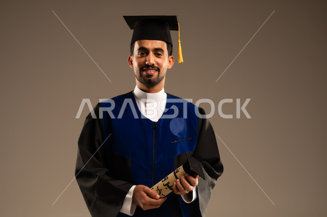 النظر الى الكاميرا بايماءات تدل على الشعور بالفخر والسعاده, بورتريه مقرب لطالب عربي سعودي خليجي يرتدي العباءه وقبعة التخرج يبتسم يمسك بيده شهادة التفوق والنجاح, الاحتفال بإنهاء الدراسة الجامعية، خلفية بنية