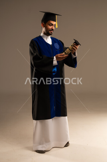 ايماءات تدل على الشعور بالفخر والسعاده, بورتريه لطالب عربي سعودي خليجي يرتدي العباءه وقبعة التخرج يمسك بيده شهادة التفوق والنجاح و ينظر اليها, الاحتفال بإنهاء الدراسة الجامعية، صورة الجسم بالطول الكامل, خلفية بنية