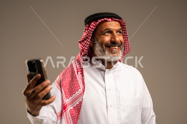  التواصل والاطمئنان على الأصدقاء, تعابير الفرح عند سماع الاخبار السارة, بورتريه مقرب لرجل مسن عربي خليجي سعودي مبتسم يرتدي العباءة والشماغ يستعمل الجوال و ينظر الى شيء ما، تصفح الانترنت والتسوق الالكتروني، خلفية بنية
