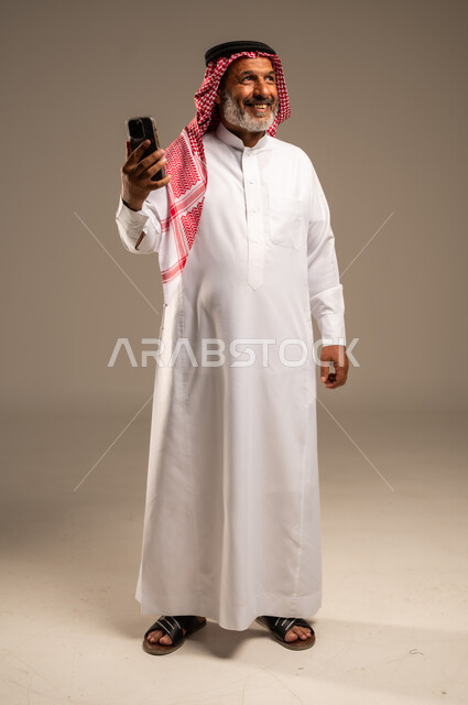  التواصل والاطمئنان على الأصدقاء, تعابير الفرح عند سماع الاخبار السارة, بورتريه لرجل مسن عربي خليجي سعودي مبتسم يرتدي العباءة والشماغ يستعمل الجوال و ينظر الى شيء ما، تصفح الانترنت والتسوق الالكتروني، الطول الكامل للجسم، خلفية بنية