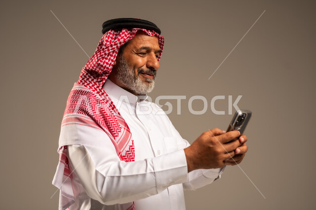  التواصل مع الافراد عبر الهاتف المحمول، دردشة ومحادثات وتعارف، بورتريه مقرب من الجانب لرجل مسن عربي خليجي سعودي مبتسم يرتدي العباءة و الشماغ يستعمل الجوال وينظر اليه بايماءات السرور والفرح، تصفح الانترنت والتسوق الالكتروني، خلفية بنية 