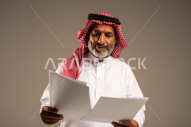 متابعة المراسلات والسجلات، بورتريه مقرب لرجل مسن عربي خليجي سعودي يرتدي العباءة والشماغ مبتسم يحمل بيديه اوراق بيضاء و ينظر اليهم، انجاز المهام المطلوبة, خلفية بنية 