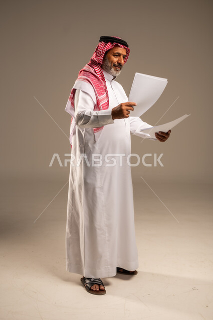 انجاز المهام المطلوبة, متابعة المراسلات والسجلات، بورتريه لرجل مسن عربي خليجي سعودي يرتدي العباءة والشماغ مبتسم يحمل بيديه اوراق بيضاء و ينظر اليهم، الطول الكامل للجسم، خلفية بنية