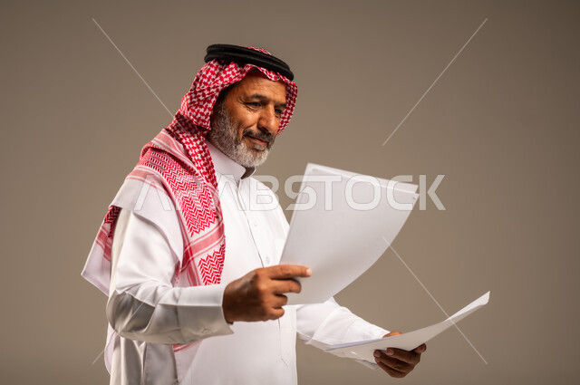 انجاز المهام المطلوبة, متابعة المراسلات والسجلات، بورتريه مقرب لرجل مسن عربي خليجي سعودي يرتدي العباءة والشماغ مبتسم يحمل بيديه اوراق بيضاء و ينظر اليهم، خلفية بنية 