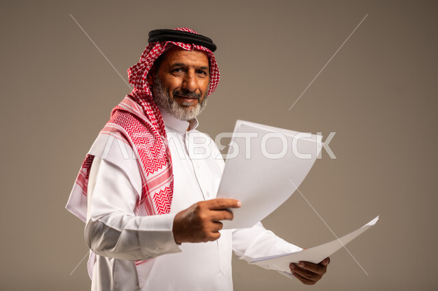 انجاز المهام المطلوبة, متابعة المراسلات والسجلات، بورتريه مقرب لرجل مسن عربي خليجي سعودي يرتدي العباءة والشماغ مبتسم يحمل بيديه اوراق بيضاء و ينظر الى الكاميرا، خلفية بنية 