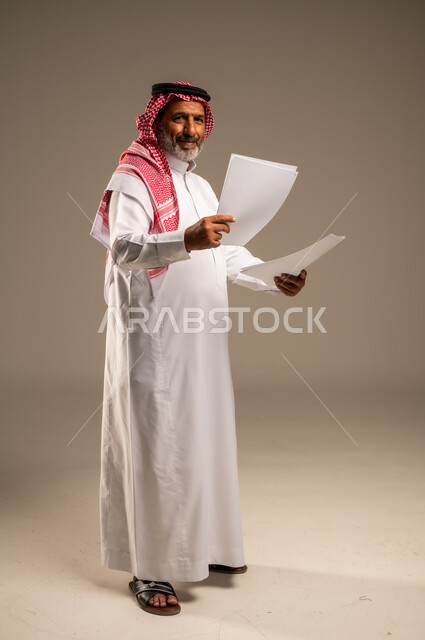 انجاز المهام المطلوبة, متابعة المراسلات والسجلات، بورتريه  لرجل مسن عربي خليجي سعودي يرتدي العباءة والشماغ مبتسم يحمل بيديه اوراق بيضاء و ينظر الى الكاميرا، الطول الكامل للجسم، خلفية بنية 