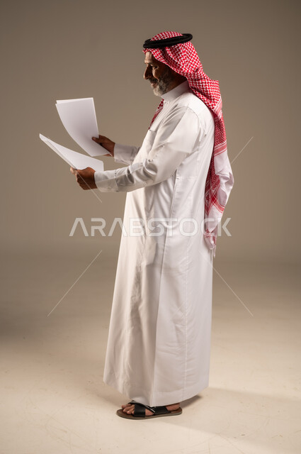 متابعة المراسلات والسجلات، بورتريه من الجانب لرجل مسن عربي خليجي سعودي يرتدي العباءة والشماغ يحمل بيديه اوراق بيضاء و ينظر اليهم، انجاز المهام المطلوبة, الطول الكامل للجسم، خلفية بنية 