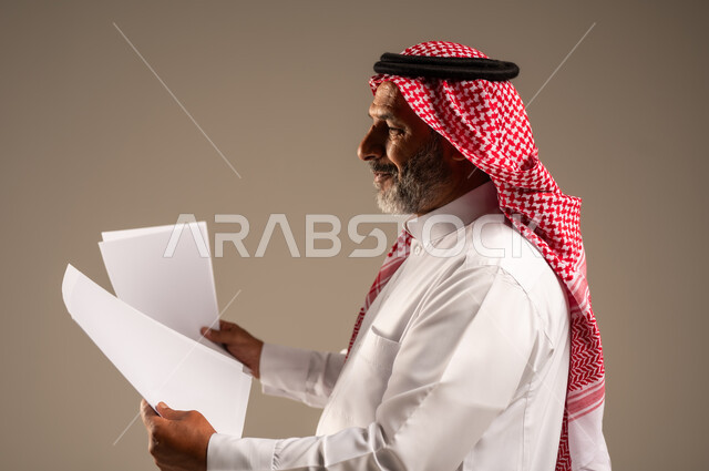 Following up correspondence and records, close-up side portrait of an elderly Saudi Arabian Gulf man wearing an abaya and shemagh holding white papers in his hands and looking at them, completing the required tasks, brown background