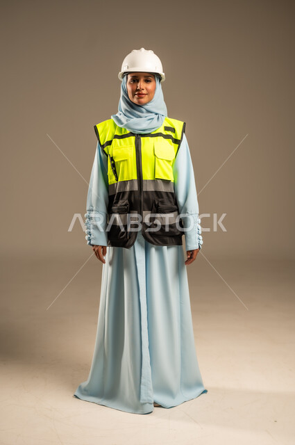مهن ووظائف نسائية هندسية, مفهوم الهندسة و الإنشاءات, العمل في المجال الهندسي, بورتريه لمهندسة عربية خليجية سعودية مبتسمة ترتدي الحجاب والخوذة وسترة الحماية تقف باستقامة تنظر إلى الكاميرا بتعابير السرور، صورة الجسم بالطول الكامل, خلفية رمادية