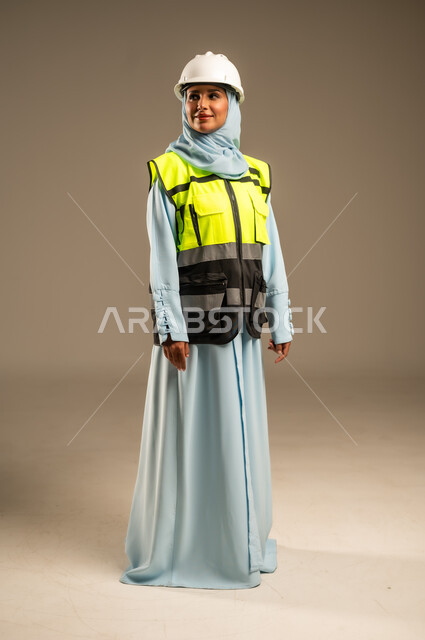 Engineering and construction concept, female engineering professions and jobs, working in the engineering field, portrait of a smiling Saudi Arabian Gulf female engineer wearing a hijab, helmet and protective vest standing straight looking at something with an expression of pleasure, full body image, gray background