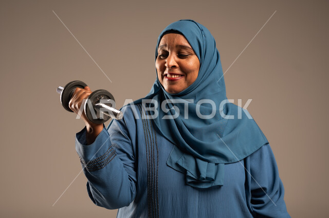 ممارسة التمارين الرياضية ورفع الاثقال مع الدمبل، بورتريه مقرب لامرآة عربية خليجية سعودية محجبة مبتسمة ترتدي العباءة تحمل بيدها الأوزان الحديدية الثقيلة، المحافظة على الصحة واللياقة الجسدية، خلفية بيج 