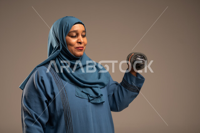 ممارسة التمارين الرياضية ورفع الاثقال مع الدمبل، بورتريه مقرب لامرآة عربية خليجية سعودية محجبة مبتسمة ترتدي العباءة تحمل بيدها الأوزان الحديدية الثقيلة، المحافظة على الصحة واللياقة الجسدية، خلفية بيج 