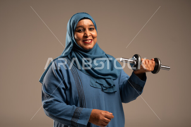 ممارسة التمارين الرياضية ورفع الاثقال مع الدمبل، بورتريه مقرب لامرآة عربية خليجية سعودية محجبة مبتسمة ترتدي العباءة تنظر الى الكاميرا تحمل بيدها الأوزان الحديدية الثقيلة، المحافظة على الصحة واللياقة الجسدية، خلفية بيج 