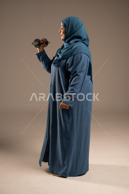 ممارسة التمارين الرياضية ورفع الاثقال مع الدمبل، بورتريه من الجانب لامرآة عربية خليجية سعودية محجبة مبتسمة ترتدي العباءة تحمل بيدها الأوزان الحديدية الثقيلة، المحافظة على الصحة واللياقة الجسدية، الجسم بالطول الكامل, خلفية بيج 