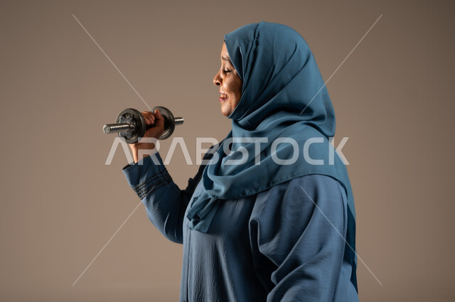 ممارسة التمارين الرياضية ورفع الاثقال مع الدمبل، بورتريه مقرب من الجانب لامرآة عربية خليجية سعودية محجبة مبتسمة ترتدي العباءة تحمل بيدها الأوزان الحديدية الثقيلة، المحافظة على الصحة واللياقة الجسدية، خلفية بيج 