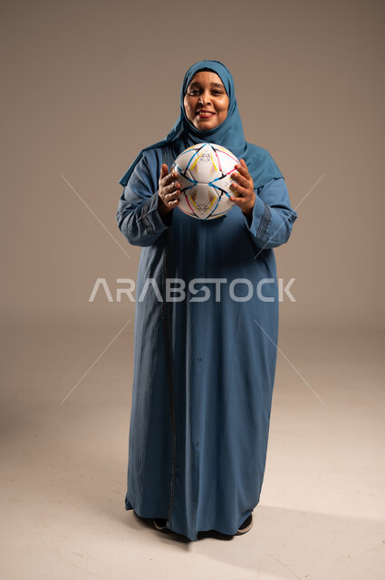 الحماس والتفاعل مع المنتخب، إيماءات الشغف والتشجيع للفريق، بورتريه لامرآة عربية خليجية سعودية محجبة مبتسمة تنظر الى الكاميرا ترتدي العباءة تحمل بيدها كرة القدم، أنديه رياضية مختلفة، الجسم بالطول الكامل, خلفية بيج