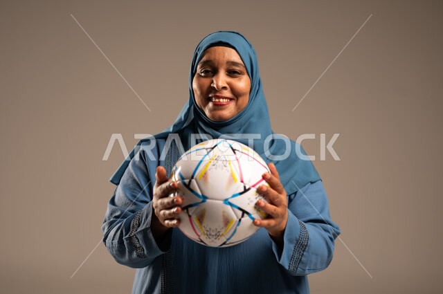 الحماس والتفاعل مع المنتخب، إيماءات الشغف والتشجيع للفريق، بورتريه مقرب لامرآة عربية خليجية سعودية محجبة مبتسمة تنظر الى الكاميرا ترتدي العباءة تحمل بيدها كرة القدم، أنديه رياضية مختلفة، خلفية بيج