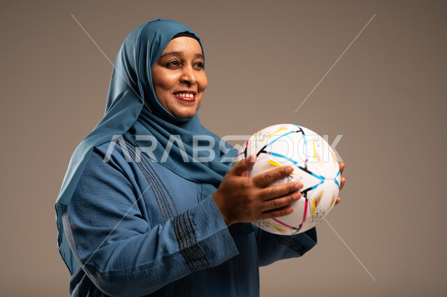 الحماس والتفاعل مع المنتخب، إيماءات الشغف والتشجيع للفريق، بورتريه مقرب لامرآة عربية خليجية سعودية محجبة مبتسمة ترتدي العباءة تحمل بيدها كرة القدم، أنديه رياضية مختلفة، خلفية بيج