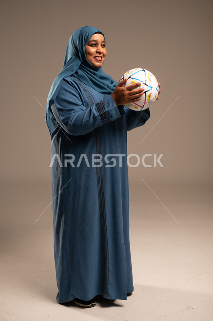 أنديه رياضية مختلفة، الحماس والتفاعل مع المنتخب، إيماءات الشغف والتشجيع للفريق، بورتريه من الجانب لامرآة عربية خليجية سعودية محجبة مبتسمة ترتدي العباءة تحمل بيدها كرة القدم، خلفية بيج