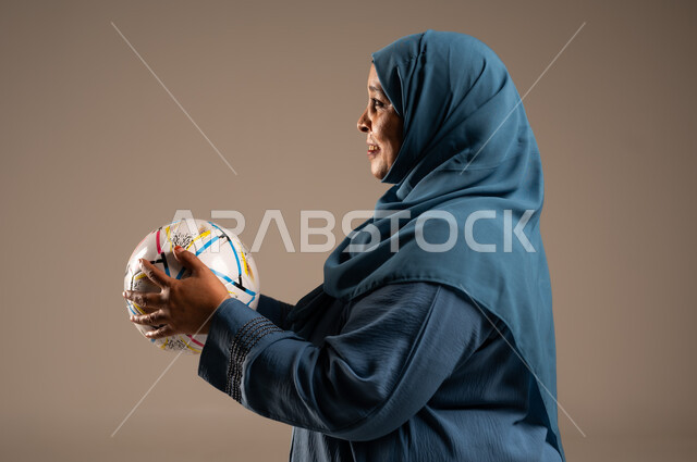 أنديه رياضية مختلفة، الحماس والتفاعل مع المنتخب، إيماءات الشغف والتشجيع للفريق، بورتريه مقرب من الجانب لامرآة عربية خليجية سعودية محجبة مبتسمة ترتدي العباءة تحمل بيدها كرة القدم، خلفية بيج