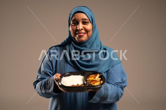 مبادرات وتبرعات انسانية خيرية، الاستمتاع بتناول الوجبات السريعة, بورتريه مقرب لأمراة كبيرة بالسن عربية خليجية سعودية عربية مبتسمة ترتدي العباءة تنظر الى الكاميرا بإيماءات الفرح تمسك بيدها وجبة طعام، توفير الغذاء للمحتاجين، خلفية بيج 