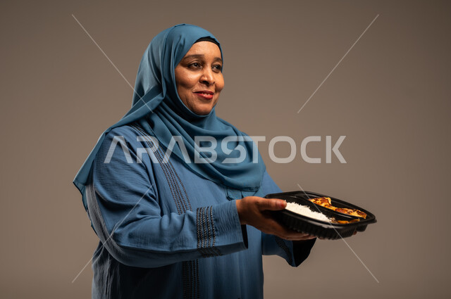 الاستمتاع بتناول الوجبات السريعة, مبادرات وتبرعات انسانية خيرية، بورتريه مقرب لأمراة كبيرة بالسن عربية خليجية سعودية عربية مبتسمة ترتدي العباءة تمسك بيدها وجبة طعام، توفير الغذاء للمحتاجين، خلفية بيج 
