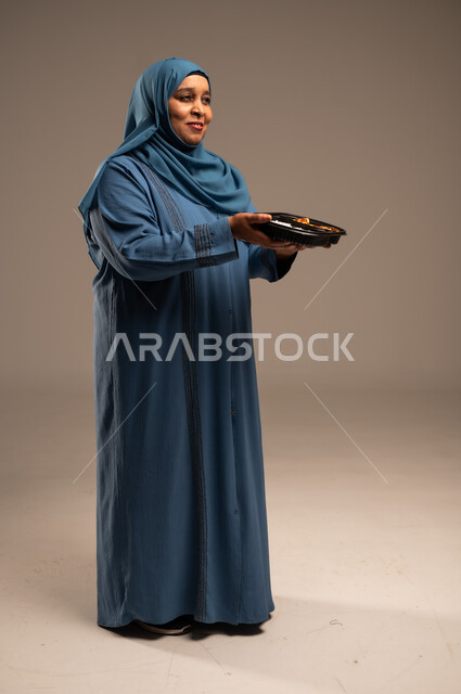 مبادرات وتبرعات انسانية خيرية، الاستمتاع بتناول الوجبات السريعة, بورتريه لأمراة كبيرة بالسن عربية خليجية سعودية عربية مبتسمة ترتدي العباءة تمسك بيدها وجبة طعام، توفير الغذاء للمحتاجين، صورة الجسم بالطول الكامل، خلفية بيج 