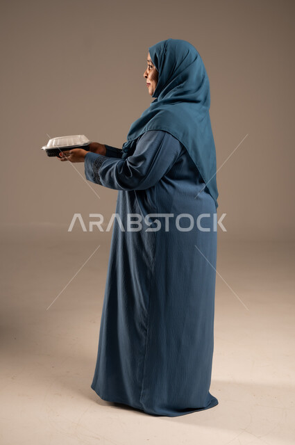 مبادرات وتبرعات انسانية خيرية، الاستمتاع بتناول الوجبات السريعة, بورتريه من الجانب لأمراة كبيرة بالسن عربية خليجية سعودية عربية مبتسمة ترتدي العباءة تمسك بيدها وجبة طعام، توفير الغذاء للمحتاجين، صورة الجسم بالطول الكامل، خلفية بيج 
