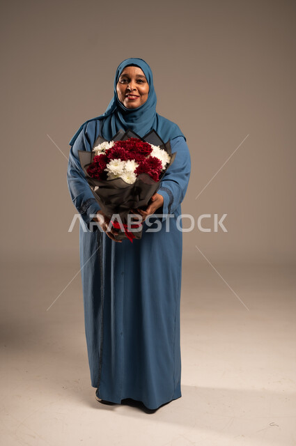 تقديم الازهار بالمناسبات، ايماءات وجه تدل على الحب، بورتريه لإمرأة كبيرة بالسن عربية خليجية سعودية محجبة ترتدي العباءة تمسك بيدها بوكيه ورد احمر، تنظر الى الكاميرا بتعابير الفرح والسرور، الجسم بالطول الكامل، خلفية بيج 