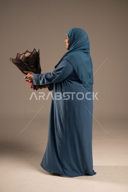 ايماءات وجه تدل على الحب، بورتريه من الجانب لإمرأة كبيرة بالسن عربية خليجية سعودية محجبة ترتدي العباءة تمسك بيدها بوكيه ورد احمر، تعابير فرح وسرور، تقديم الازهار بالمناسبات، الجسم بالطول الكامل، خلفية بيج 