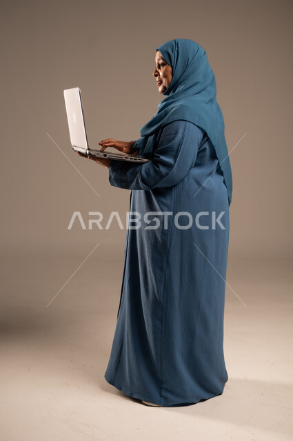 استخدام الأجهزة الحديثة والمتطورة، ادارة الأعمال عن بعد، بورتريه من الجانب لامرأة كبيرة بالسن عربية خليجية سعودية ترتدي العباءة والحجاب تقوم باستخدام الحاسب المحمول، دمج التكنولوجيا بالعمل، إنجاز المهام المطلوبة عبر اللابتوب، الجسم بالطول الكامل، خلفية بيج