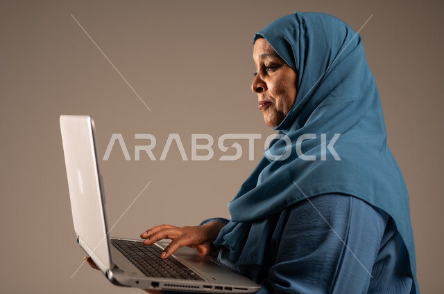 استخدام الأجهزة الحديثة والمتطورة، ادارة الأعمال عن بعد، بورتريه مقرب من الجانب لامرأة كبيرة بالسن عربية خليجية سعودية ترتدي العباءة والحجاب تقوم باستخدام الحاسب المحمول، دمج التكنولوجيا بالعمل، إنجاز المهام المطلوبة عبر اللابتوب، خلفية بيج
