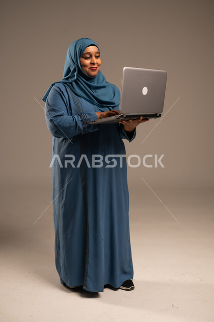 استخدام الأجهزة الحديثة والمتطورة، ادارة الأعمال عن بعد، بورتريه لامرأة كبيرة بالسن عربية خليجية سعودية ترتدي العباءة والحجاب تقوم باستخدام الحاسب المحمول، دمج التكنولوجيا بالعمل، إنجاز المهام المطلوبة عبر اللابتوب، الجسم بالطول الكامل، خلفية بيج