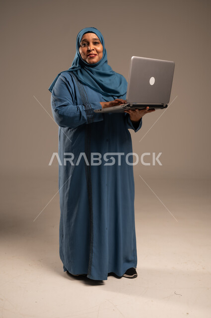 ادارة الأعمال عن بعد، استخدام الأجهزة الحديثة والمتطورة، بورتريه لامرأة كبيرة بالسن عربية خليجية سعودية ترتدي العباءة والحجاب تنظر الى الكاميرا، دمج التكنولوجيا بالعمل، إنجاز المهام المطلوبة عبر اللابتوب، مهن ووظائف نسائية، الجسم بالطول الكامل، خلفية بيج
