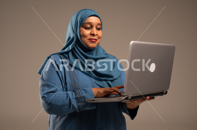 ادارة الأعمال عن بعد، استخدام الأجهزة الحديثة والمتطورة، بورتريه مقرب لامرأة كبيرة بالسن عربية خليجية سعودية ترتدي العباءة والحجاب تقوم باستخدام الحاسب المحمول، دمج التكنولوجيا بالعمل، إنجاز المهام المطلوبة عبر اللابتوب، مهن ووظائف نسائية، خلفية ملونة  