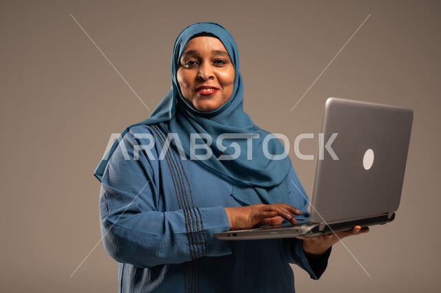 ادارة الأعمال عن بعد، استخدام الأجهزة الحديثة والمتطورة، بورتريه مقرب لامرأة كبيرة بالسن عربية خليجية سعودية ترتدي العباءة والحجاب تنظر الى الكاميرا، دمج التكنولوجيا بالعمل، إنجاز المهام المطلوبة عبر اللابتوب، مهن ووظائف نسائية، خلفية ملونة  