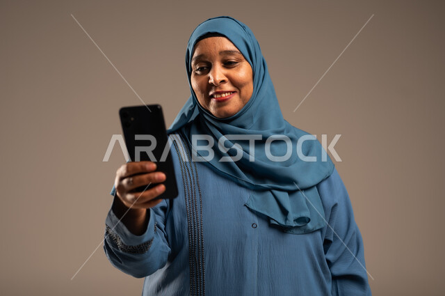 تبادل الأحاديث مع الأهل والأصدقاء، تصفح التطبيقات وبرامج التواصل الاجتماعي، بورتريه مقرب لإمرأة كبيرة بالسن عربية خليجية سعودية محجبة مبتسمة ترتدي العباءة تمسك بيدها الجوال و تنظر اليه، استخدام الأجهزة والتقنيات الحديثة والمتطورة، خلفية بيج 