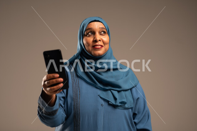 تبادل الأحاديث مع الأهل والأصدقاء، تصفح التطبيقات وبرامج التواصل الاجتماعي، بورتريه مقرب لإمرأة كبيرة بالسن عربية خليجية سعودية محجبة مبتسمة ترتدي العباءة تمسك بيدها الجوال تنظر الى شيء ما، استخدام الأجهزة والتقنيات الحديثة والمتطورة، خلفية بيج 
