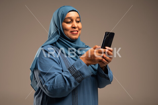 تصفح التطبيقات وبرامج التواصل الاجتماعي، تبادل الأحاديث مع الأهل والأصدقاء، بورتريه مقرب لإمرأة كبيرة بالسن عربية خليجية سعودية محجبة مبتسمة ترتدي العباءة تمسك بيدها الجوال، استخدام الأجهزة والتقنيات الحديثة والمتطورة، خلفية بيج 