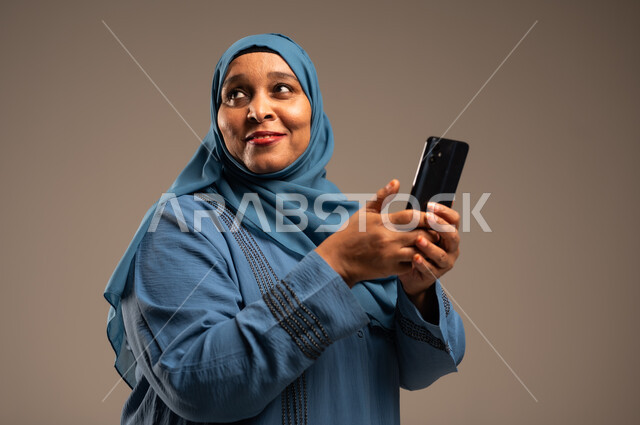 تصفح التطبيقات وبرامج التواصل الاجتماعي، تبادل الأحاديث مع الأهل والأصدقاء، بورتريه مقرب لإمرأة كبيرة بالسن عربية خليجية سعودية محجبة ترتدي العباءة تمسك بيدها الجوال تنظر الى شيء ما، استخدام الأجهزة والتقنيات الحديثة والمتطورة، خلفية بيج 