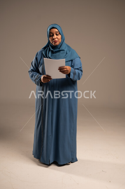 متابعة المراسلات والسجلات، بورتريه لامرأة كبيرة بالسن عربية خليجية سعودية ترتدي العباءة والحجاب تحمل بيديها اوراق بيضاء و تنظر اليهم بتعابير الجدية، انجاز المهام المطلوبة, الطول الكامل للجسم، خلفية بنية 