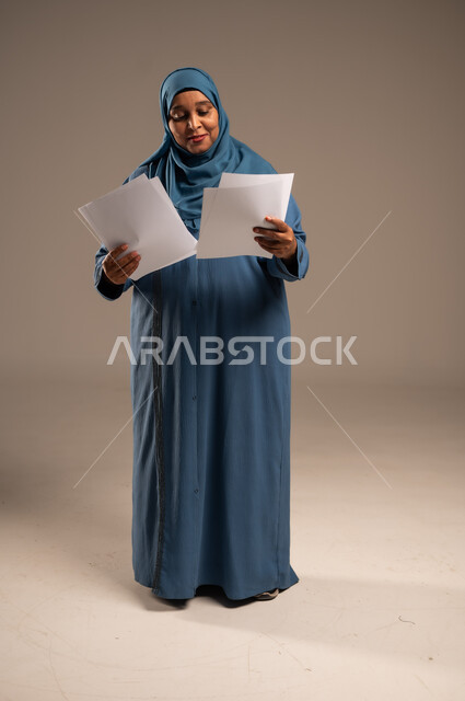 متابعة المراسلات والسجلات، بورتريه لامرأة كبيرة بالسن عربية خليجية سعودية ترتدي العباءة والحجاب تحمل بيديها اوراق بيضاء و تنظر اليهم بتعابير الفرح، انجاز المهام المطلوبة, الطول الكامل للجسم، خلفية بنية 
