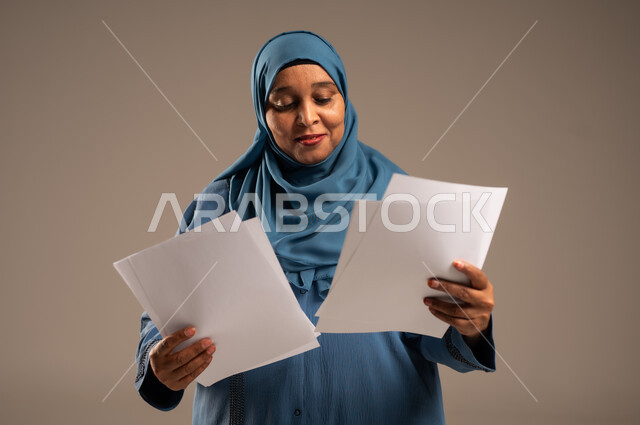 انجاز المهام المطلوبة, متابعة المراسلات والسجلات، بورتريه مقرب لامرأة كبيرة بالسن عربية خليجية سعودية ترتدي العباءة والحجاب مبتسمة تحمل بيديها اوراق بيضاء و تنظر اليهم، خلفية بنية 