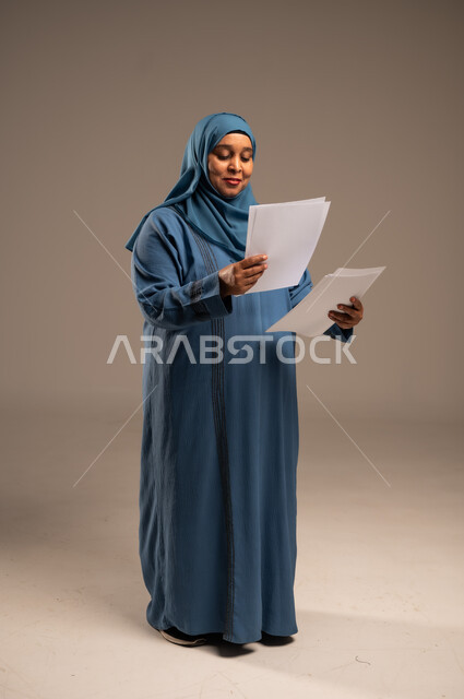متابعة المراسلات والسجلات، بورتريه لامرأة كبيرة بالسن عربية خليجية سعودية ترتدي العباءة والحجاب تحمل بيديها اوراق بيضاء و تنظر اليهم، انجاز المهام المطلوبة, الطول الكامل للجسم، خلفية بنية 