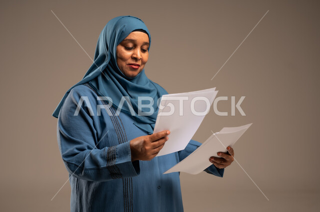 انجاز المهام المطلوبة, متابعة المراسلات والسجلات، بورتريه مقرب لامرأة كبيرة بالسن عربية خليجية سعودية ترتدي العباءة والحجاب مبتسمة تحمل بيديها اوراق بيضاء و تنظر اليهم، خلفية بنية 