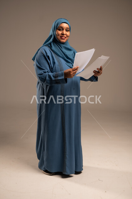 انجاز المهام المطلوبة, متابعة المراسلات والسجلات، بورتريه لامرأة كبيرة بالسن عربية خليجية سعودية ترتدي العباءة والحجاب مبتسمة تحمل بيديها اوراق بيضاء و تنظر الى الكاميرا، الطول الكامل للجسم، خلفية بنية 