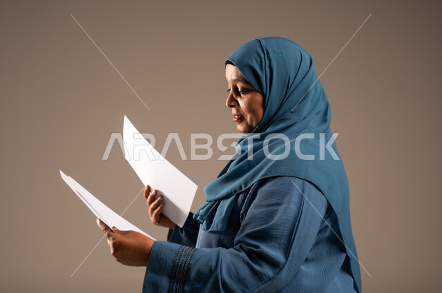 انجاز المهام المطلوبة, متابعة المراسلات والسجلات، بورتريه مقرب من الجانب لامرأة كبيرة بالسن عربية خليجية سعودية ترتدي العباءة والحجاب مبتسمة تحمل بيديها اوراق بيضاء و تنظر اليهم، خلفية بنية 