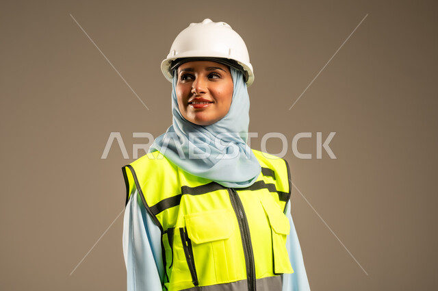 Working in the engineering field, close-up portrait of a smiling Saudi Arabian Gulf female engineer wearing hijab, helmet and safety vest standing straight looking at something with an expression of pleasure, engineering and construction concept, female engineering professions and jobs, gray background