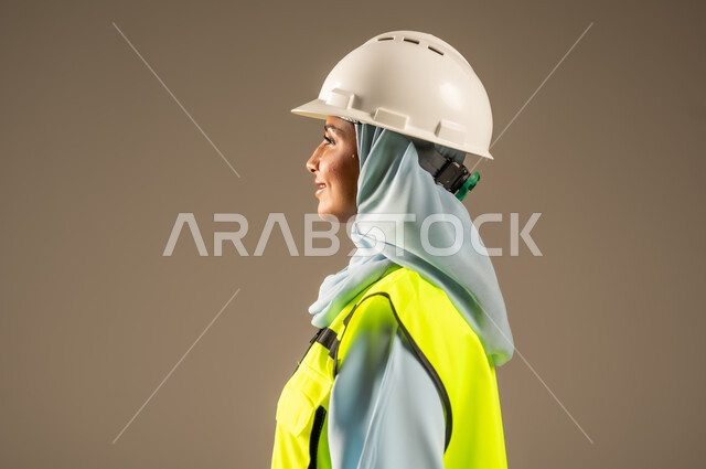 العمل في المجال الهندسي, بورتريه مقرب من الجانب لمهندسة عربية خليجية سعودية مبتسمة ترتدي الحجاب والخوذة وسترة الحماية تقف باستقامة تنظر إلى شيء ما بتعابير السرور، مفهوم الهندسة و الإنشاءات, مهن ووظائف نسائية هندسية, خلفية رمادية