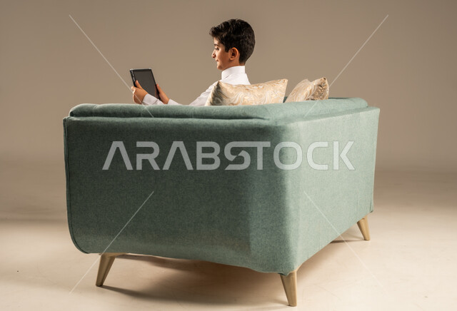 استخدام الأجهزة التقنية الحديثة، بورتريه من الخلف لطفل عربي سعودي خليجي يرتدي الثوب التقليدي يجلس على كرسي مريح ويقوم باستخدام التابلت، مفهوم التعليم وإنجاز المهام عن بعد، قضاء وقت ممتع على الإنترنت، اللعب على الجهاز اللوحي، خلفية بيج